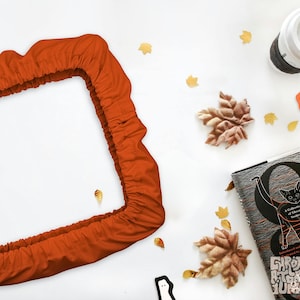 Peut inclure: Un tissu orange de forme rectangulaire, possiblement une taie d'oreiller ou une housse, est au centre de l'image. Des feuilles d'automne décoratives, une tasse à café et un livre avec une illustration de chat sont dispersés. Thème automnal.