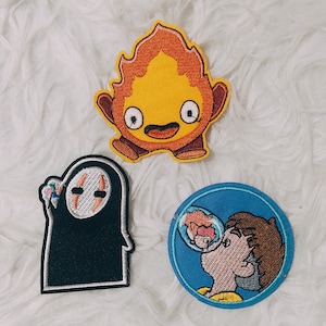 Peut inclure: Trois patchs brodés thermocollants représentant des personnages du film d'animation 'Le Voyage de Chihiro'. Le premier patch est un esprit de feu jaune et orange, le deuxième est un esprit sans visage noir et blanc, et le troisième est un personnage bleu et brun soufflant une bulle.