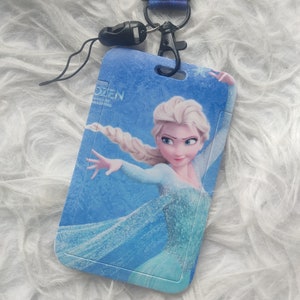 Puede incluir: Un cordón azul con un cierre negro y un tarjetero con Elsa de Frozen. El tarjetero tiene el número 10 impreso. El cordón está sobre una superficie blanca y esponjosa.