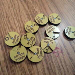 Puede incluir: Un conjunto de fichas de acción amarillas con bordes negros, cada una con la inscripción "ACTION TOKEN". Las fichas presentan un diseño de diagrama circular con un pequeño remache plateado en el centro. Las fichas están dispersas sobre una superficie de madera.
