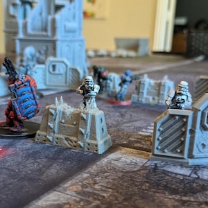 May include: A tabletop wargaming scene with miniature figures and terrain pieces. The figures are painted in white, red, and blue. The terrain pieces are made of gray plastic and feature walls and barricades.