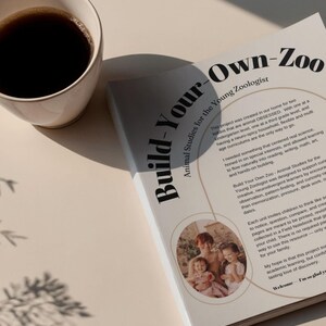 May include: A white mug filled with dark coffee sits next to a book titled "Build-Your-Own-Zoo: Animal Studies for the Young Zoologist." The book features a family photo and text. The scene is lit with soft, natural light.