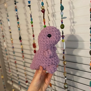 May include: A soft, purple crocheted dinosaur toy with black eyes. The toy is being held by a hand in front of a white background with colorful beaded strings hanging down.