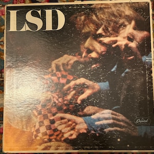 Könnte beinhalten: Ein Vintage-Vinyl-Schallplattencover mit dem Titel "LSD" in fetten weißen Buchstaben. Das Cover-Artwork zeigt eine Nahaufnahme einer Gruppe von Händen, die ein kariertes Muster halten, mit einem verschwommenen Bild eines Gesichts im Hintergrund. Das Capitol Records-Logo befindet sich in der unteren rechten Ecke.