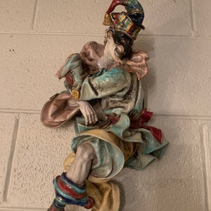 Op de afbeelding: Een keramische wandsculptuur van een nar die een veelkleurige hoed en een masker draagt. De nar is geschilderd in tinten blauw, groen, geel, rood en wit.