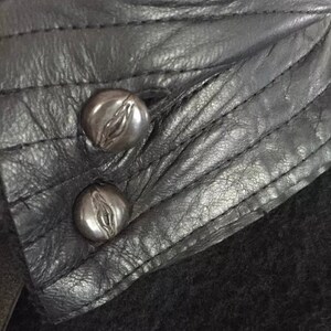 May include: Close-up of a black leather jacket collar with two silver metal buttons. The buttons have a decorative design.
