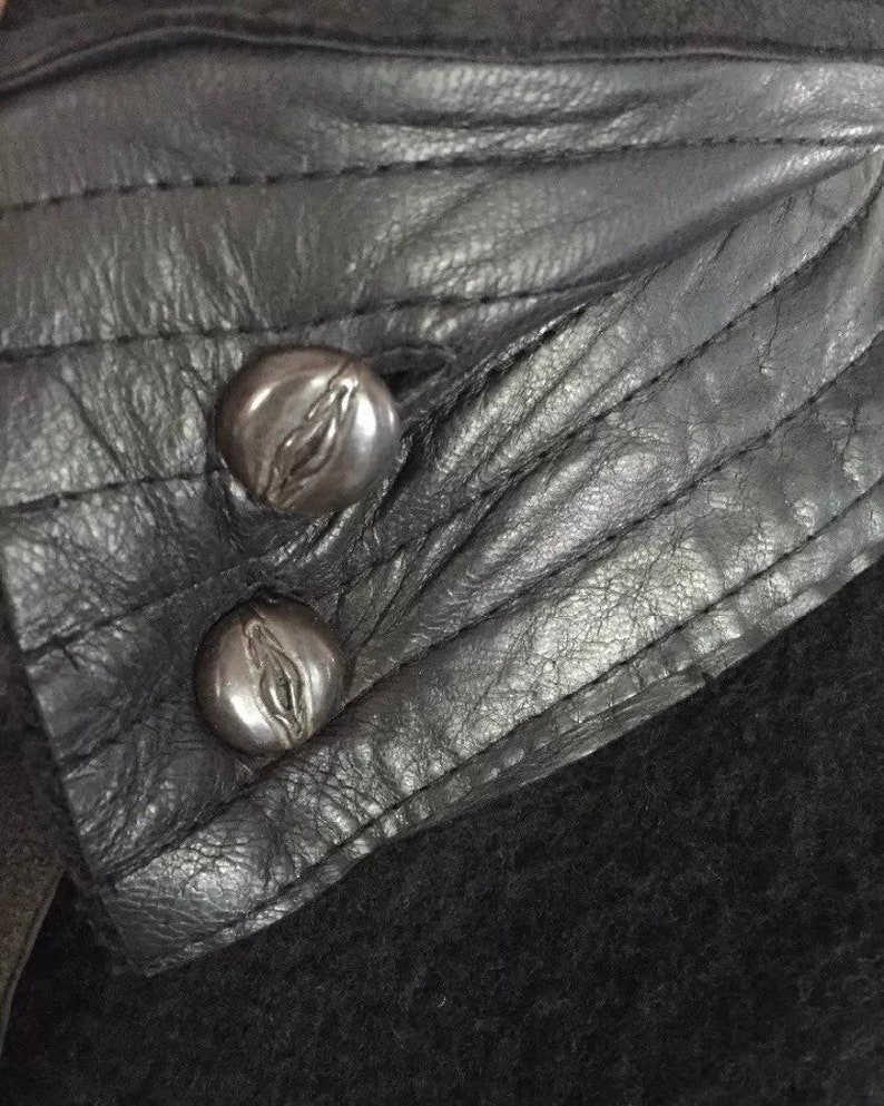 May include: Close-up of a black leather jacket collar with two silver metal buttons. The buttons have a decorative design.