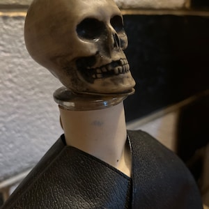 May include: A black leather bottle holder with a skull-shaped stopper. The skull is made of a light brown material and has a detailed face.