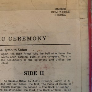 May include: A close-up of a record album cover with the title "Side II" and text about the Satanic Bible by Anton Szandor LaVey. The text describes the four books that make up the Satanic Bible, including The Book of Satan, The Book of Lucifer, The Book of Belial, and The Book of Leviathan.