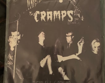 May include: Black and white photo of a band called "The Cramps" on a record sleeve. The text "Human Fly / Domino" is printed below the band's name.