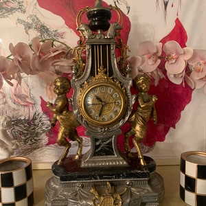 May include: A decorative clock with a gold and silver finish, featuring two cherub figures and a black marble base. The clock face has Roman numerals and a gold trim.