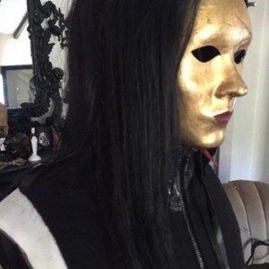 May include: A person wearing a black jacket with a zip and a gold mask covering their face. The mask has eye holes and a painted mouth.