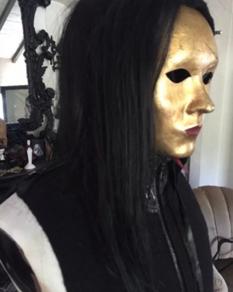 May include: A person wearing a black jacket with a zip and a gold mask covering their face. The mask has eye holes and a painted mouth.