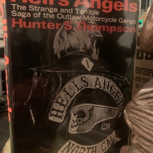 May include: A black and white book cover with the title "Hell's Angels: The Strange and Terrible Saga of the Outlaw Motorcycle Gangs" by Hunter S. Thompson. The cover features a black and white image of a biker wearing a leather jacket with a "Hells Angels" patch on the back.