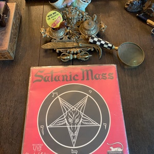 May include: A red vinyl record with a black pentagram and the text "Satanic Mass" in white. The record is labeled "Recorded Live at the Church of Satan, San Francisco" and "Morgenstrom Stereo".