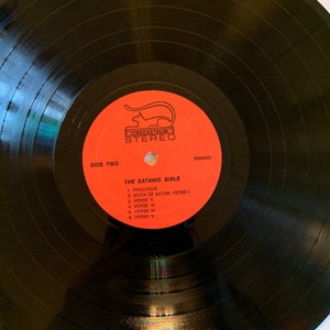 May include: Black vinyl record with a red label. The label has a cat logo and the text "MORGENSTRUM STEREO" and "THE SATANIC BIBLE". The track list includes "PROLOGUE", "BOOK OF SATAN, VERSE I", "VERSE II", "VERSE III", "VERSE IV", and "VERSE V".