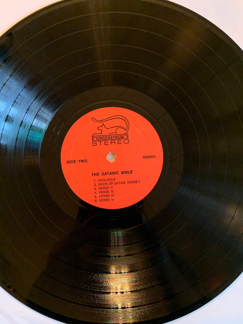 May include: Black vinyl record with a red label. The label has a cat logo and the text "MORGENSTRUM STEREO" and "THE SATANIC BIBLE". The track list includes "PROLOGUE", "BOOK OF SATAN, VERSE I", "VERSE II", "VERSE III", "VERSE IV", and "VERSE V".