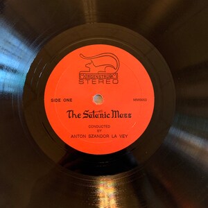 May include: A black vinyl record with a red label. The label has a black and white illustration of a cat, the text "Murgenstrum Stereo", and the title "The Satanic Mass" with the text "Conducted by Anton Szandor La Vey".