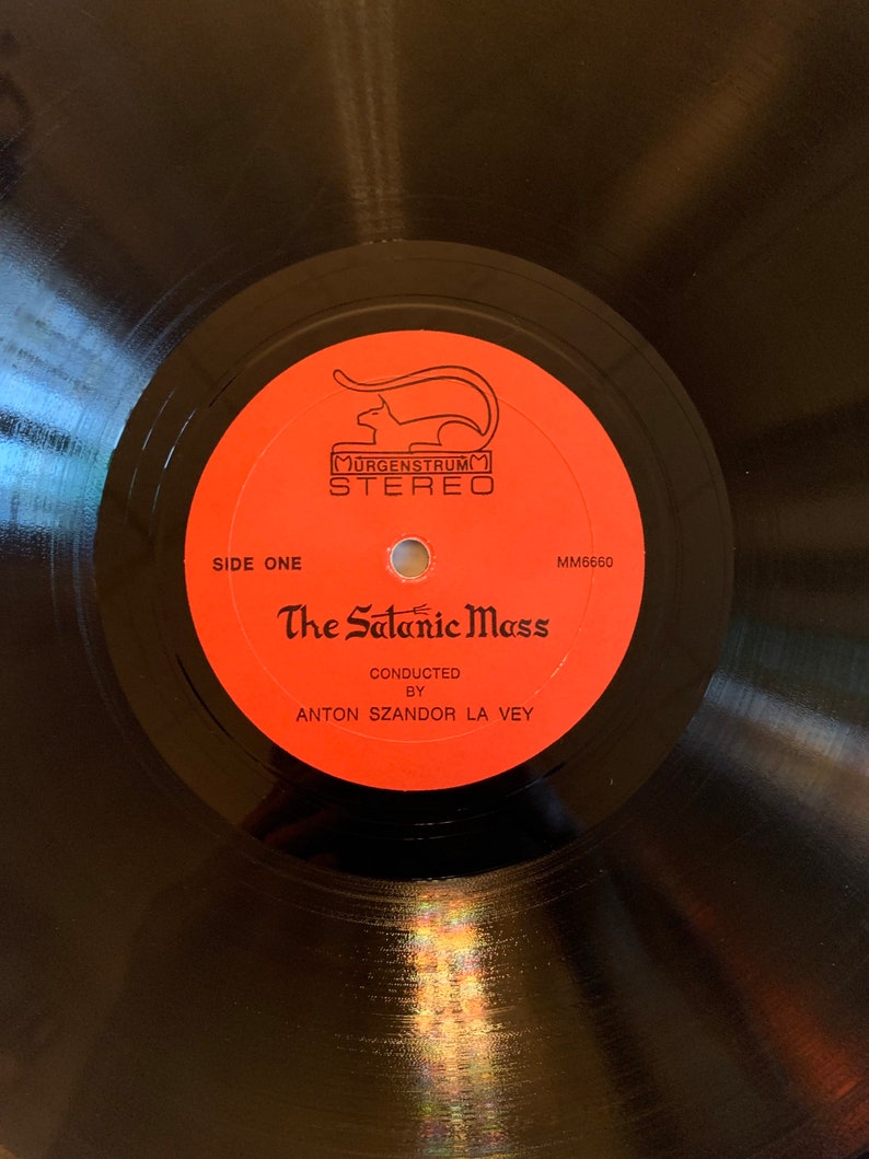 May include: A black vinyl record with a red label. The label has a black and white illustration of a cat, the text "Murgenstrum Stereo", and the title "The Satanic Mass" with the text "Conducted by Anton Szandor La Vey".