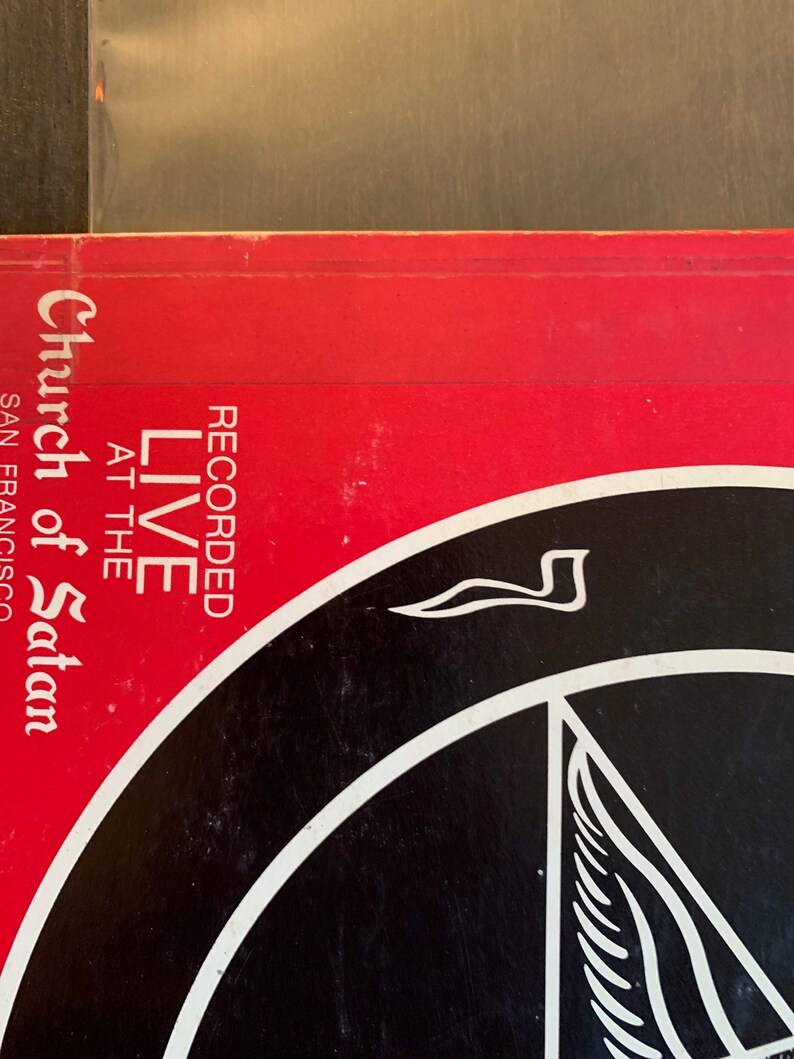 May include: A red and black vinyl record cover with the text "Recorded Live at the Church of Satan San Francisco" and a black and white pentagram with a winged symbol.