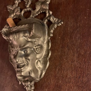 May include: A vintage metal wall mounted ashtray with a decorative design of a face and grape vines. The ashtray has a small compartment for holding cigarettes.