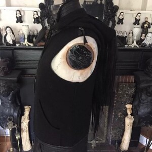 May include: Black leather vest with a large eye detail on the front. The vest has a long black fringe hanging from the back.