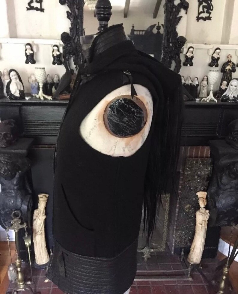 May include: Black leather vest with a large eye detail on the front. The vest has a long black fringe hanging from the back.