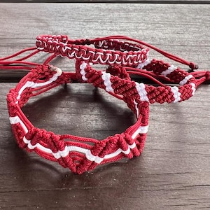 May include: Three handmade macrame bracelets in red and white. The bracelets are made with a knotted design, featuring a combination of red and white cords. The bracelets are displayed on a wooden surface.
