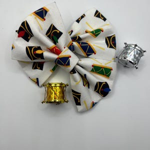 Puede incluir: Un lazo de tela blanco con un patrón de tambores y sombreros de juguete rojos, azules, verdes y amarillos. Dos pequeños tambores de juguete, uno dorado y otro plateado, están al lado del lazo.