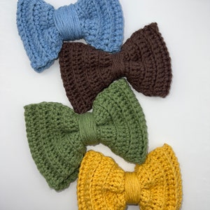 May include: Four crocheted bows in different colors: light blue, brown, green, and yellow. The bows are arranged in a stack, with the light blue bow on top and the yellow bow on the bottom.