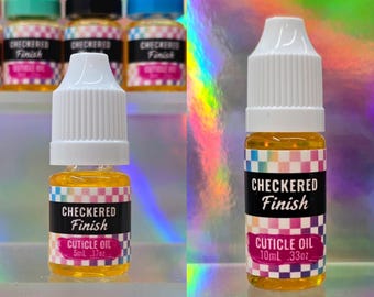 Puede incluir: Dos frascos de plástico transparente con aceite para cutículas amarillo y tapones de rosca blancos. Las etiquetas presentan un patrón de cuadros de colores, el texto "CHECKERED Finish" en blanco y "CUTICLE OIL" con 5ml y 10ml.