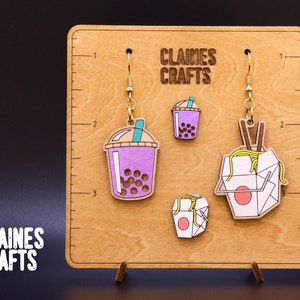 May include: A pair of wooden earrings featuring a purple boba tea and a yellow takeout container with chopsticks. The earrings are displayed on a wooden board with the text "Claines Crafts" printed on it.