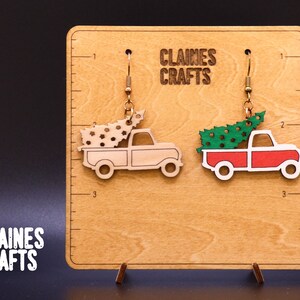 May include: A pair of Christmas tree earrings featuring a red truck with a green tree and a white truck with a brown tree. The earrings are displayed on a wooden board with the text "Claines Crafts".