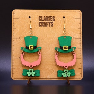 May include: St Patrick's Day earrings made of wood, featuring a green leprechaun hat with a black band and gold buckle, a pink beard, and a green bow tie with a shamrock. The earrings hang from gold-coloured hooks. The earrings are displayed on a wooden board with the text "CLAINE'S CRAFTS".