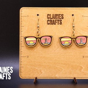 Puede incluir: Un par de pendientes de madera con forma de gafas de sol con un atardecer y un faro pintados en las lentes. Los pendientes cuelgan de ganchos dorados en una tarjeta de exhibición de madera con el texto "Claines Crafts".