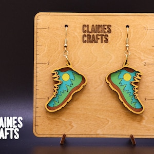 May include: A pair of wooden earrings shaped like hiking boots with a green, blue, and yellow mountain scene. The earrings are hanging from gold hooks on a wooden display with the text "Claines Crafts".
