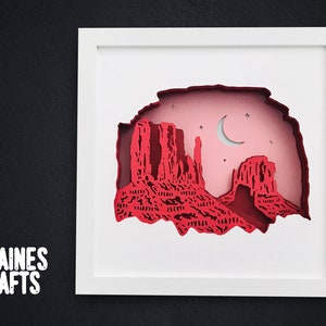 Puede incluir: Una obra de arte en papel enmarcada en blanco que representa un paisaje desértico rojo con una luna creciente y estrellas en un cielo rosa. El título "Clainies Crafts" es visible en la esquina inferior izquierda.