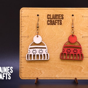 May include: A pair of wooden earrings shaped like winter hats. The earrings are displayed on a wooden board with the text "Claines Crafts". One earring is red with white snowflakes and the other is white with red snowflakes.