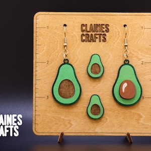 May include: A wooden display board with a ruler on the side shows four avocado-shaped earrings. The earrings are green with brown centers and have gold hooks. The board has the text "Claines Crafts" printed on it.