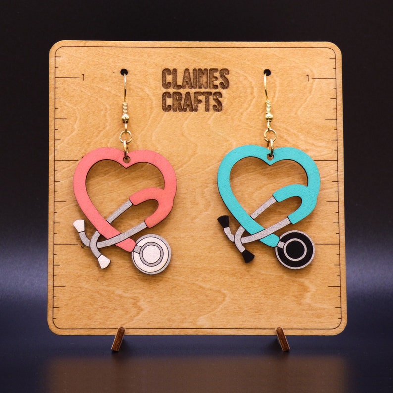 May include: A pair of wooden earrings shaped like hearts with a stethoscope inside. The earrings are on a wooden display stand with the text "Claines Crafts".