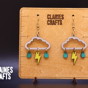 May include: A pair of wooden earrings featuring a gray cloud with yellow lightning bolt and teal raindrops hanging from it. The earrings are on a wooden display with the text "Claines Crafts".
