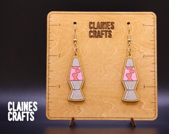 May include: A pair of wooden earrings shaped like lava lamps. The earrings are painted in shades of pink, gray, and white. The earrings are hanging from a wooden display board with the text "Claines Crafts" printed on it.