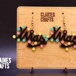 Puede incluir: Un par de pendientes negros de luces de Navidad con luces de colores. Los pendientes están hechos de madera y tienen ganchos dorados. Los pendientes se muestran en una tabla de madera con el texto "Claines Crafts" impreso en ella.