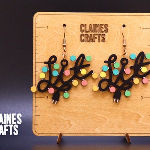 May include: A pair of black wooden earrings shaped like a string of lights with the word "Lit" in the center. The lights are colored yellow, pink, blue, and green. The earrings are hanging from gold hooks.