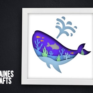May include: A white framed paper art piece depicting a blue whale with a purple body and a blue tail. The whale is surrounded by coral and fish in shades of orange, green, and blue. The whale is swimming upwards with a spray of water above it.