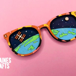 May include: A pair of wooden sunglasses with a colorful abstract design. The lenses are painted to look like a view of Earth from space, with a small space station orbiting the planet. The sunglasses are on a pink background.