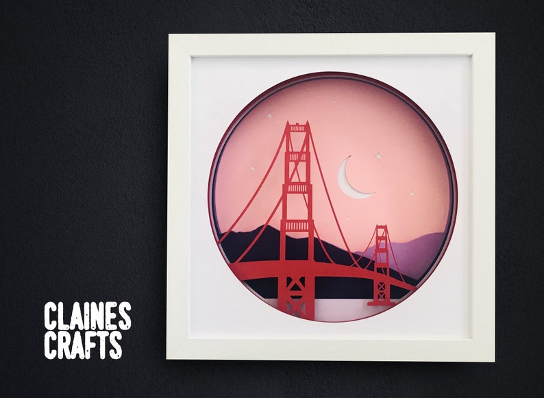 May include: A white framed paper art piece depicting the Golden Gate Bridge in a circular cutout. The bridge is red and the background is a pink sunset with a crescent moon and stars.