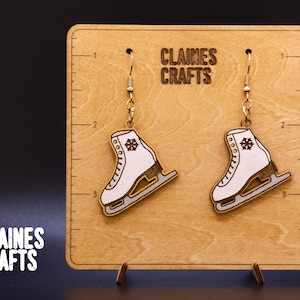 以下が含まれることがあります： 金色のアクセントと雪の結晶のデザインが施された白いスケート靴のイヤリングのペア。イヤリングは「Claines Crafts」という文字が書かれた木製のボードに飾られています。