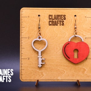 May include: A pair of wooden earrings on a display stand. The earrings are shaped like a heart with a keyhole and a key. The text "Claines Crafts" is printed on the display stand.