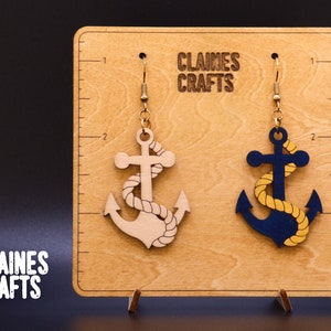 May include: A pair of wooden anchor earrings with gold hooks. The anchors are painted in blue and beige with a yellow rope design. The earrings are displayed on a wooden board with the text "Claines Crafts".